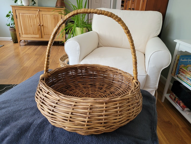 May include: A large, round, woven wicker basket with a brown handle. The basket is empty and sitting on a gray blanket.