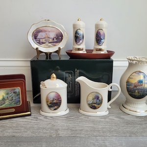 May include: A set of white porcelain kitchenware featuring images from the 'Christmas in the Country' collection. The set includes a creamer, sugar bowl, salt and pepper shakers, a napkin holder, and a decorative plate. The items are decorated with gold trim and feature various winter scenes.