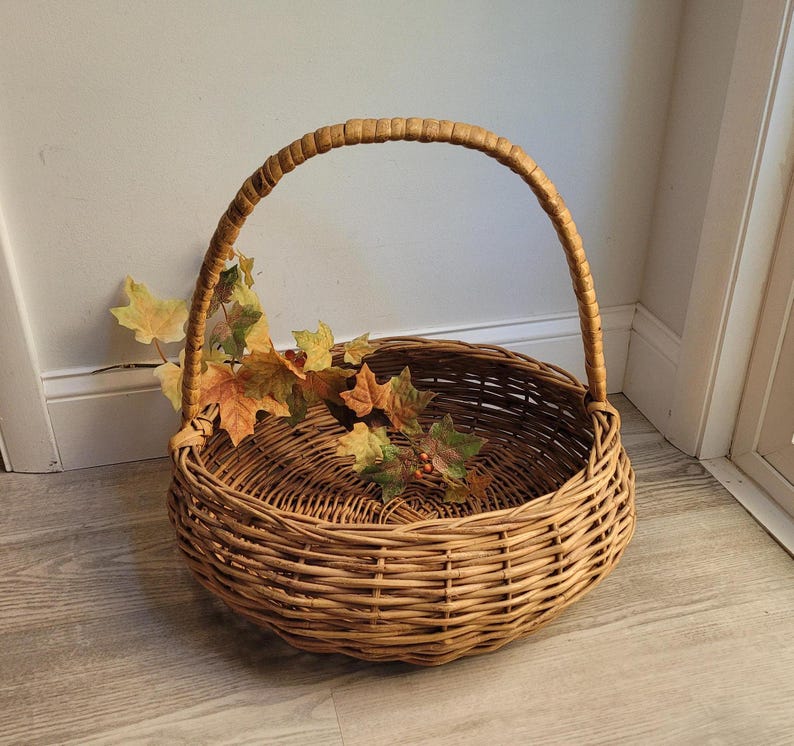 May include: A large, round wicker basket with a woven handle. The basket is filled with dried fall leaves and berries.