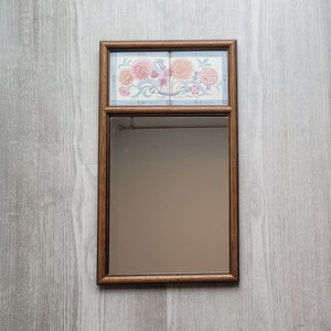 May include: A wooden framed mirror with a floral tile design in pink, blue, and white. The mirror has a brown wooden frame.