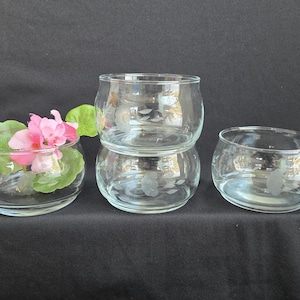 May include: Clear glass bowls, some stacked, with a round shape. One bowl contains pink flowers and green leaves. The bowls are on a dark surface, creating a contrast that highlights their transparency and form.