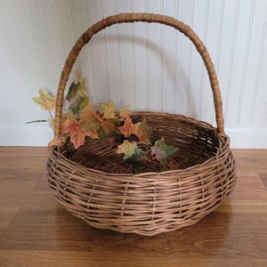 May include: A large, round wicker basket with a woven handle. The basket is filled with fall leaves and is sitting on a wooden floor.