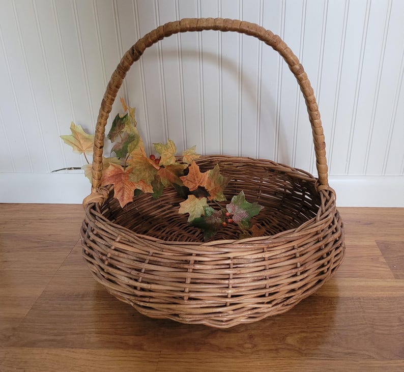 May include: A large, round wicker basket with a woven handle. The basket is filled with fall leaves and is sitting on a wooden floor.