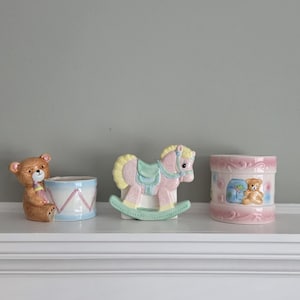 May include: Three ceramic baby-themed decorations. A brown bear figure next to a blue and white drum-shaped container. A pink, yellow, and green rocking horse. A pink container with a teddy bear illustration.
