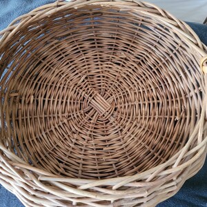 May include: A round, woven wicker basket with a handle. The basket is made of natural brown wicker and has a circular shape. The basket is empty.