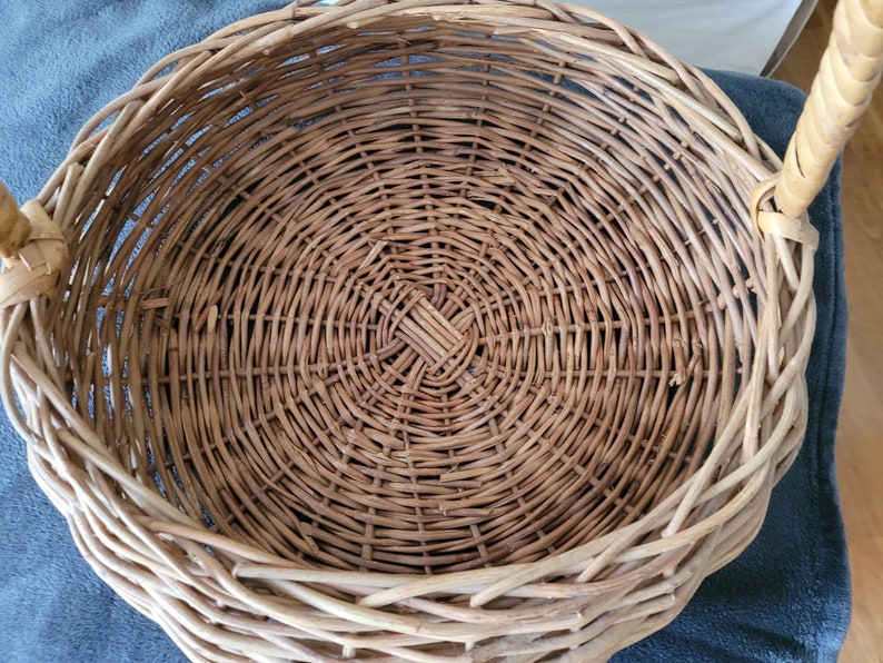 May include: A round, woven wicker basket with a handle. The basket is made of natural brown wicker and has a circular shape. The basket is empty.