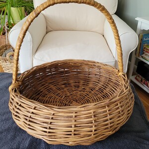 May include: A large, round wicker basket with a woven handle. The basket is made of natural brown wicker and has a wide, open top.
