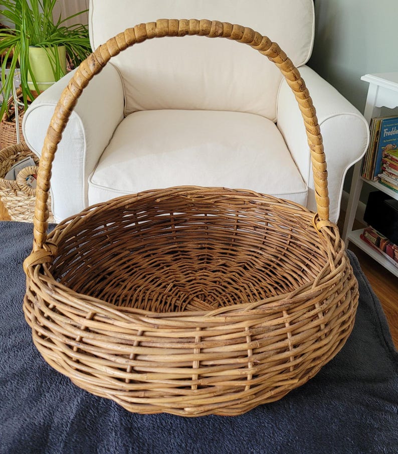 May include: A large, round wicker basket with a woven handle. The basket is made of natural brown wicker and has a wide, open top.