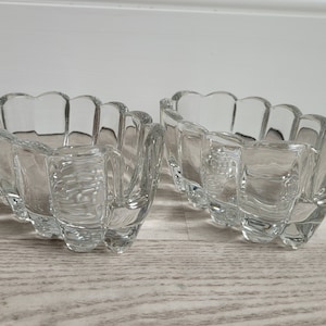 May include: Two clear glass bowls with a scalloped rim. The bowls are rectangular with a curved, petal-like design around the edges. They sit on a light-colored wooden surface.