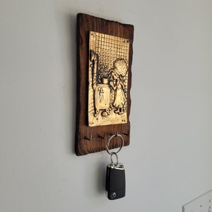 May include: A wooden key holder with a gold-toned metal plaque depicting a girl in a dress. The plaque has a grid pattern and the words "To" and "C.H." are visible. A black car key is hanging from a silver key ring on the holder.