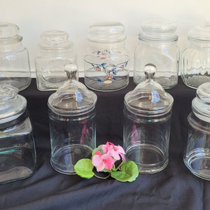 May include: Nine clear glass jars with lids. The jars are various shapes and sizes. Some of the jars have decorative designs on the lids.