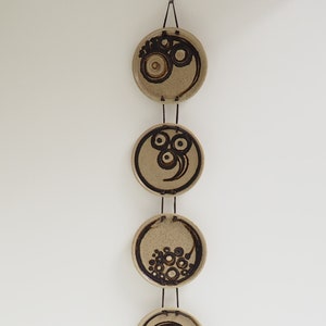 May include: A wall hanging made of five ceramic discs with black and brown abstract designs. The discs are strung together with a thin cord and hang vertically.