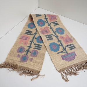 May include: A beige woven rug with a geometric pattern in pink, blue, and gray. The rug has fringe on both ends.