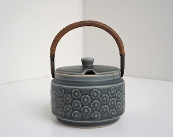 May include: A gray ceramic sugar bowl with a floral pattern and a brown wicker handle. The lid has a small spout for pouring.