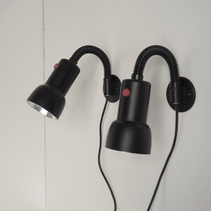 May include: Two black flexible-arm lamps with silver-coloured light bulbs mounted on a white wall. Each lamp has a red on/off switch.