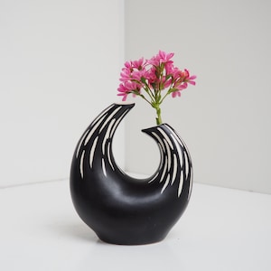 Può includere: Un vaso in ceramica nera con linee astratte bianche, a forma di mezzaluna, con fiori rosa nell'apertura.