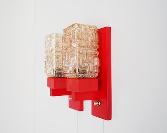 May include: A vintage wall sconce with a red wooden base and two textured glass light fixtures. The sconce has a rectangular design with a switch and a cord. The glass shades are a light amber color.