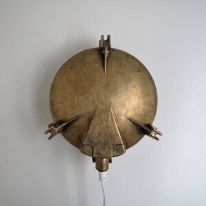 May include: A brass wall sconce with a round, slightly domed shade and a cord hanging down. The sconce has a simple, industrial design.