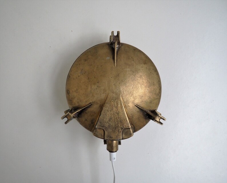 May include: A brass wall sconce with a round, slightly domed shade and a cord hanging down. The sconce has a simple, industrial design.