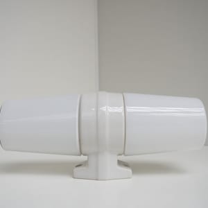 May include: White porcelain wall sconce with a cylindrical design. The sconce has a simple, modern aesthetic.