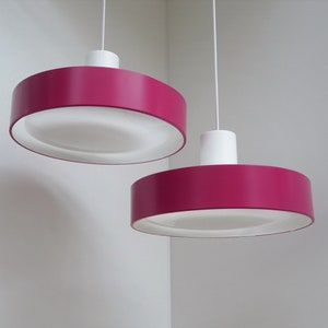 May include: Two modern pendant lights with a vibrant magenta shade and a white interior. The lights hang from white cords and have a white cylindrical top. The lamps are set against a white wall.