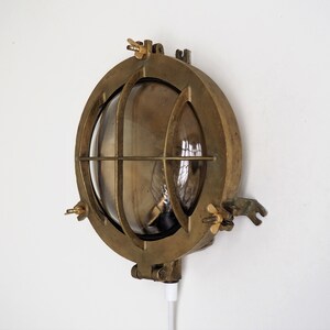 May include: A brass nautical wall light with a clear glass lens and a cage-like design. The light has a cord and plug for easy installation.