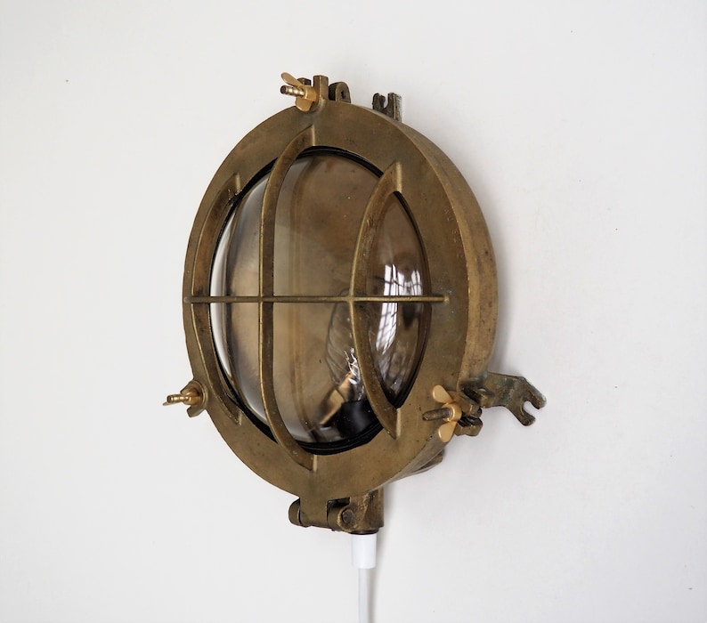 May include: A brass nautical wall light with a clear glass lens and a cage-like design. The light has a cord and plug for easy installation.