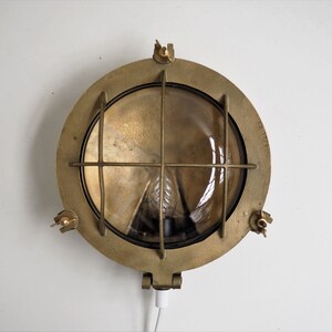 May include: A brass nautical wall light with a round, clear glass lens and a wire cage. The light has a cord and plug.