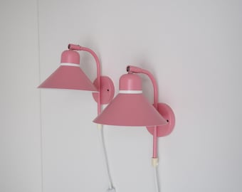 Puede incluir: Dos apliques de pared rosas a juego con una pantalla cónica y un ribete blanco. Las lámparas tienen un brazo curvo y una base redonda. Los apliques están montados en una pared blanca. Las lámparas miden unos 25 cm de alto.