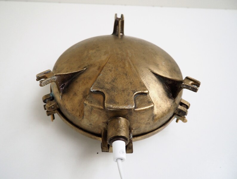 May include: A brass nautical wall light with a round, domed shade and a white cord. The light has four decorative arms that extend from the shade.