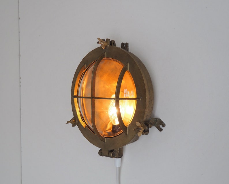 May include: A brass wall-mounted light fitting with a round, caged glass shade. The light is on and emitting a warm, yellow glow.