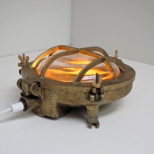 May include: A brass nautical bulkhead light with a caged glass shade. The light is turned on and emits a warm yellow glow.