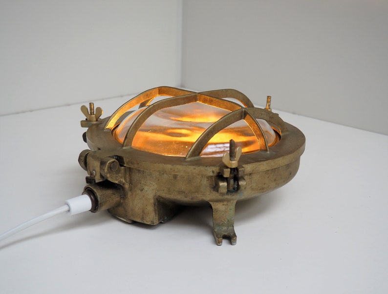 May include: A brass nautical bulkhead light with a caged glass shade. The light is turned on and emits a warm yellow glow.