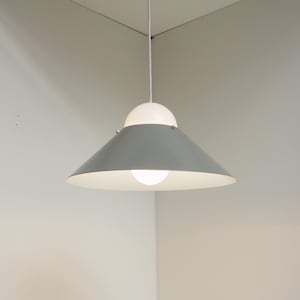 May include: A contemporary pendant light with a grey, cone-shaped shade and a white, spherical top. The light hangs from a white cord and illuminates a room corner. The light fixture has a bright, exposed bulb.