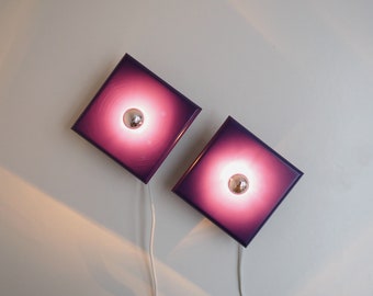 Puede incluir: Dos apliques de pared cuadrados con pantallas de vidrio morado y bombillas plateadas. Los apliques están montados en una pared blanca.