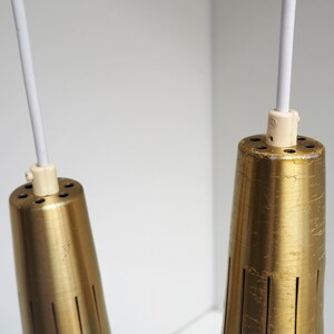 May include: Two gold-toned metal pendant lights with white cords hanging from the ceiling. The lights have a cylindrical shape with a textured surface and a series of small holes around the top.