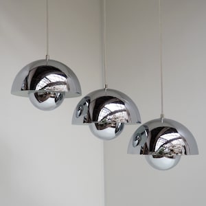 May include: Three chrome pendant lights with a reflective, dome-shaped shade and a spherical bottom. The lights hang from thin, silver cords against a white wall. The reflective surfaces mirror the surrounding environment.