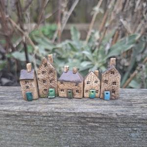 Könnte beinhalten: Eine Reihe von fünf Miniatur-Keramikhäusern mit strukturierten braunen Wänden, unterschiedlichen Dachformen und kleinen, farbigen Türen. Jedes Haus hat einen Schornstein. Die Häuser sind auf einer Holzoberfläche angeordnet.