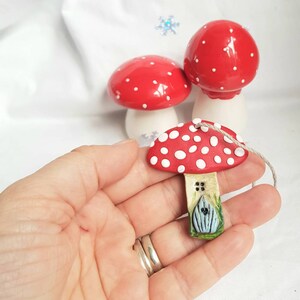 May include: A red and white ceramic mushroom ornament with a small blue door and green grass. The mushroom is hanging from a string.