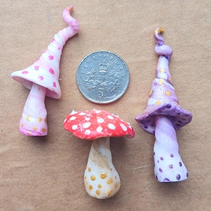 Puede incluir: Tres setas de cerámica pintadas a mano con diseños caprichosos. Una seta es roja con puntos blancos, otra es rosa con puntos dorados y la última es morada con puntos dorados.