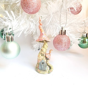 May include: A whimsical, hand-painted mushroom house ornament with a pink and yellow color scheme. The ornament features a textured design, a small door, and a spiraled top. It is displayed among Christmas tree ornaments.