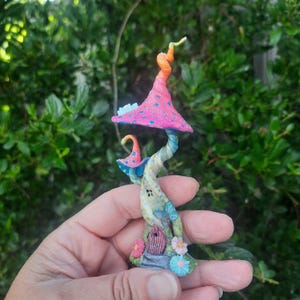 May include: A whimsical fairy house made of colourful clay, shaped like a mushroom with a pink top and a swirling, multi-coloured stem. The house has a small window and is decorated with flowers and dots.