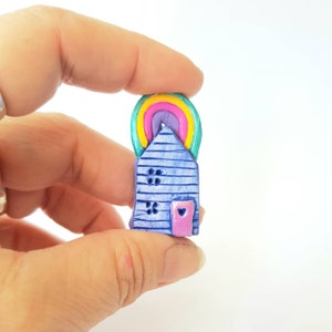 Puede incluir: Una pequeña casa azul con un arcoíris en la parte superior y una puerta rosa con un corazón. La casa está hecha de arcilla y tiene una superficie texturizada.