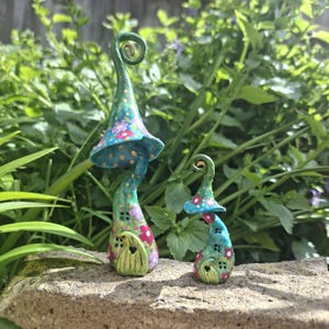 Puede incluir: Dos caprichosas casas de hadas con forma de hongo. La más grande es de color turquesa con un estampado floral en el sombrero y una pequeña puerta. La más pequeña tiene un diseño similar. Ambas tienen un tallo verde y están sobre un fondo de follaje verde.