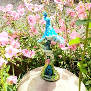 May include: A whimsical fairy house made of blue and white ceramic, shaped like a mushroom with a small door and a pink and green flower garden around the base. The house is sitting on a light brown surface.