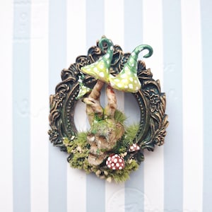 Op de afbeelding: Een handgemaakte sculptuur met een schedel, paddenstoelen en groen gestippelde hoeden in een sierlijke, antieke lijst. De schedel is omgeven door mos en kleine rood-witte paddenstoelen, wat een fantasiethema creëert.