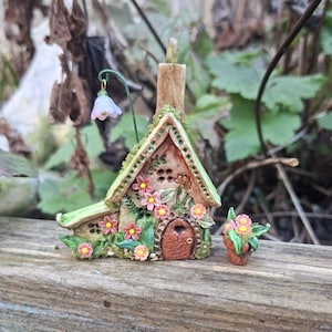 May include: A detailed miniature fairy house, featuring a brown door and pink flowers, with a small flower pot alongside. The house is adorned with green moss and a delicate hanging flower.