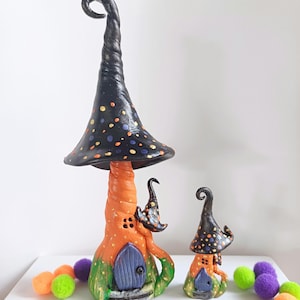 Könnte beinhalten: Zwei Halloween-Themen-Pilzhäuser aus Ton. Das größere Haus ist orange mit einem schwarzen und goldenen Tupfenhut. Das kleinere Haus ist orange mit einem schwarzen und goldenen Tupfenhut. Beide Häuser haben blaue Türen.
