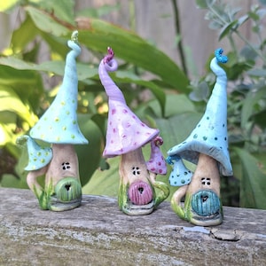 May include: Three miniature mushroom houses in pastel shades of blue, purple, and green. Each has a spiralled top, a small door, and a tiny mushroom. The houses are set against a backdrop of green foliage.