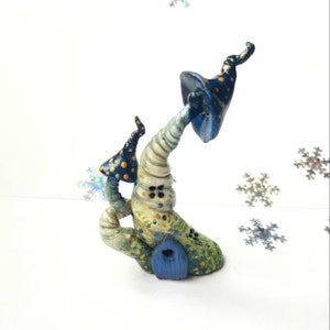 Puede incluir: Escultura de casa de hadas pintada a mano con forma de hongo. Presenta una puerta azul, un tallo texturizado y arremolinado, y sombreros de hongo azules y dorados con lunares. La base es verde con detalles azules y naranjas.